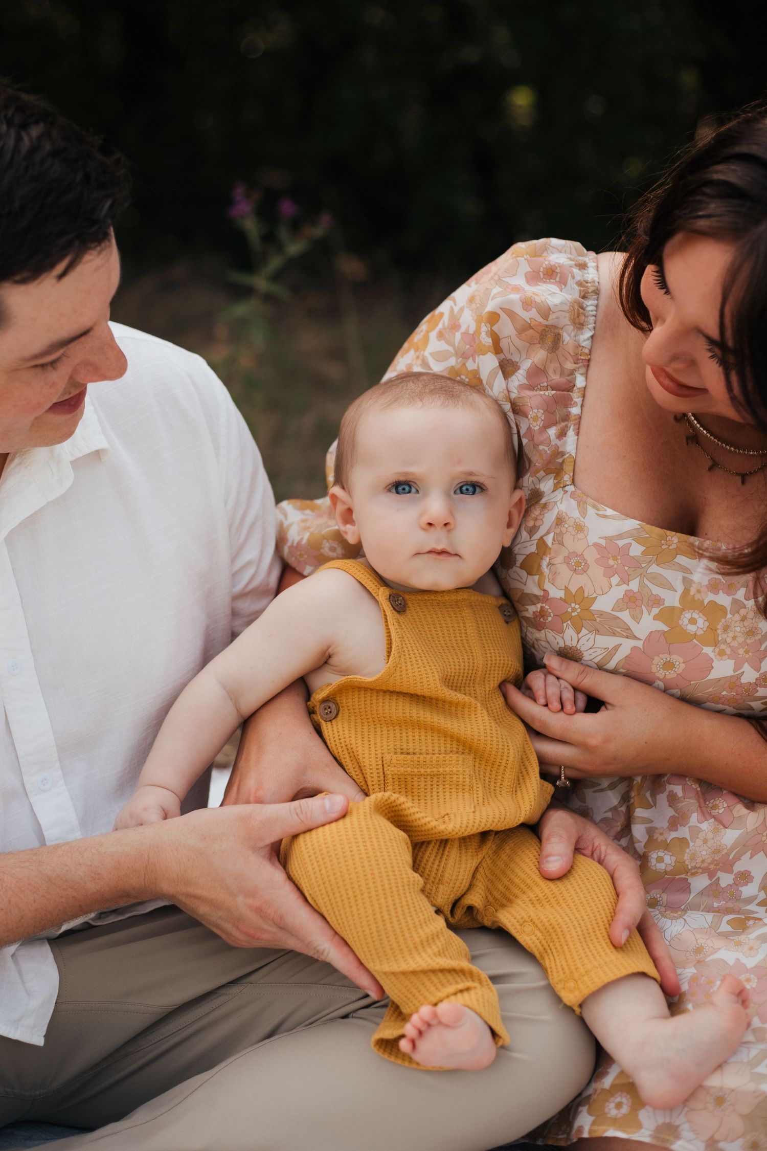 Baby in yellow romper held by parents during outdoor photoshoot.
