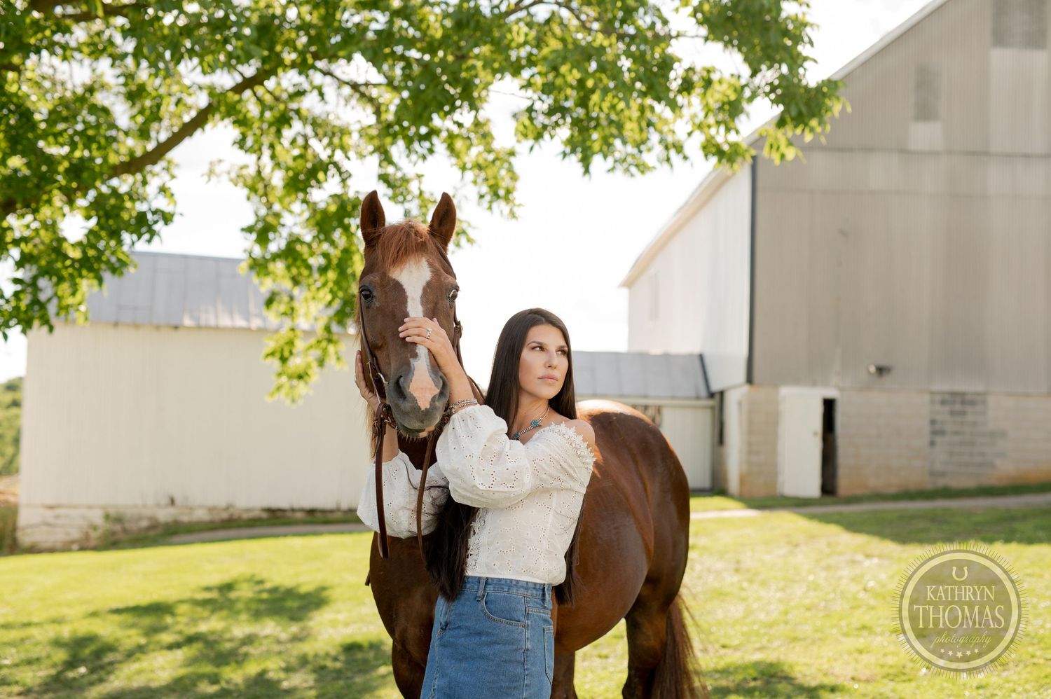 A beautiful chestnut horse and companion share a moment together near a white barn under green trees.