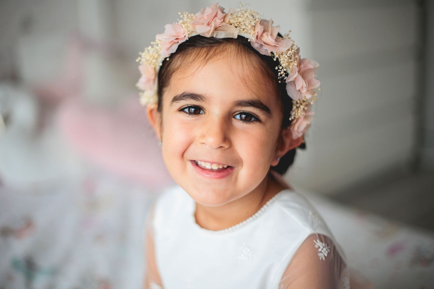 A child wearing a delicate floral crown and white dress smiles warmly in soft indoor lighting.