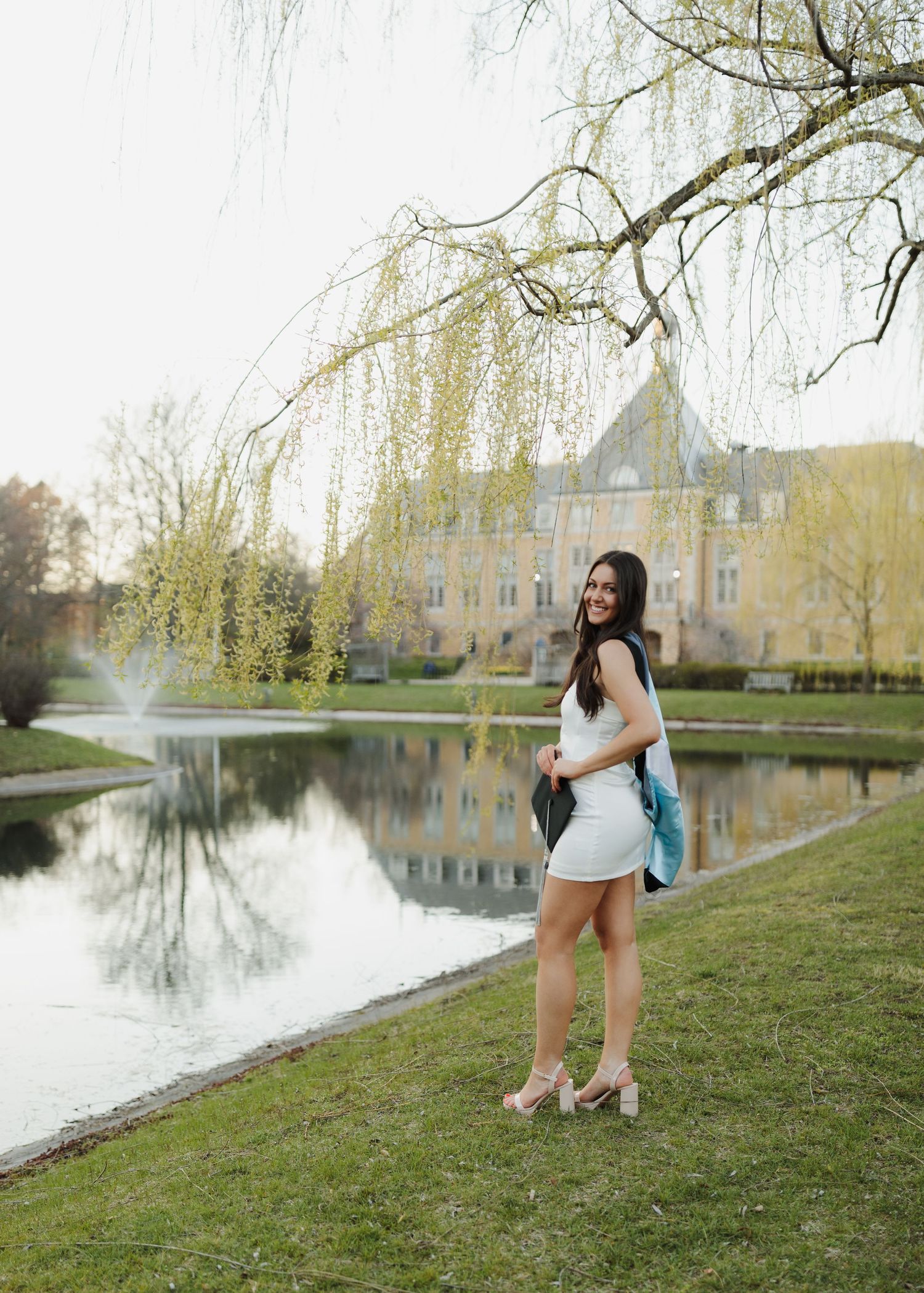 Graduate poses by reflective pond with castle-like building and willow trees in background.