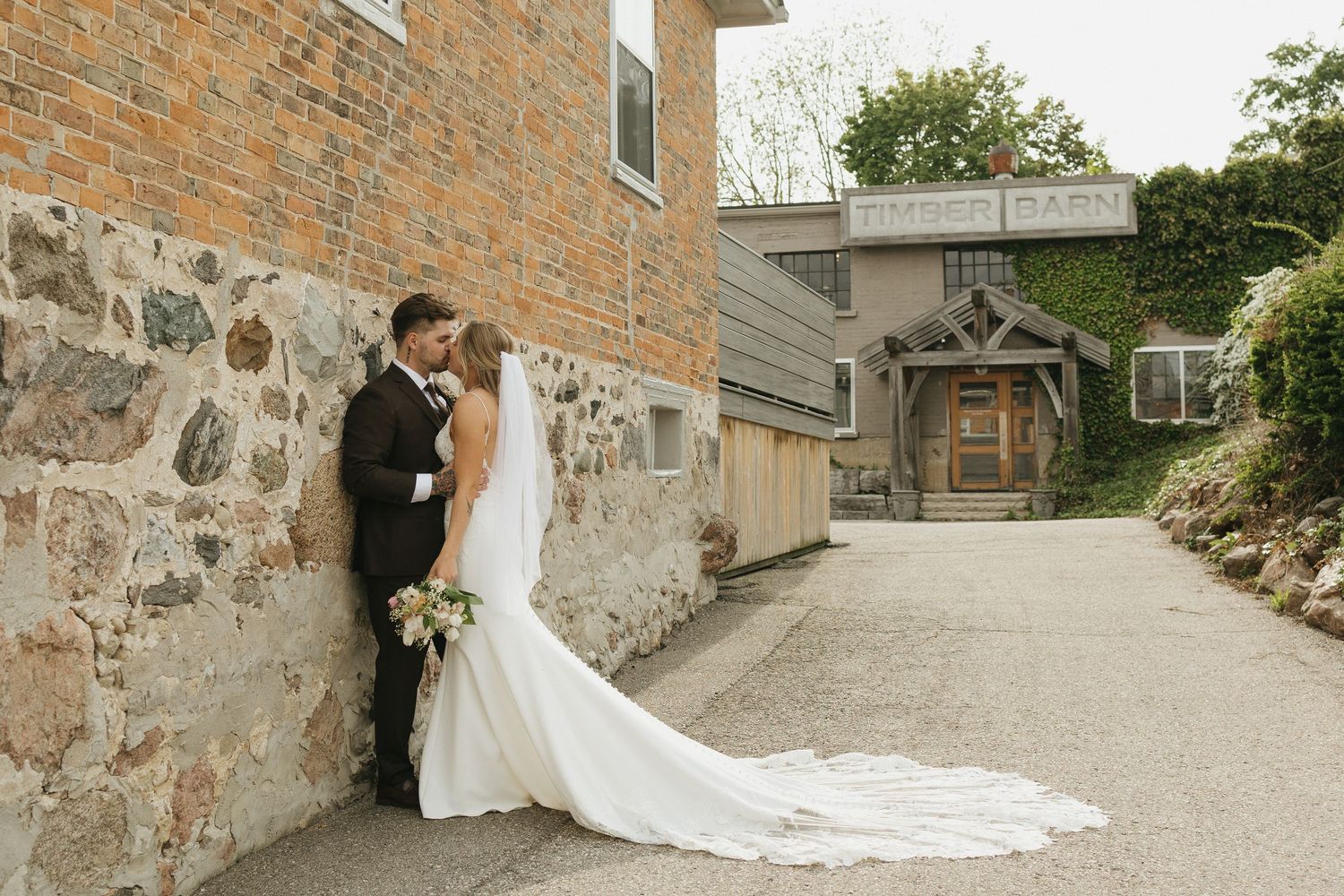 A newlywed couple leaning against a wall kissing in front of the Timberbarn wedding venue in St Jacobs Ontario