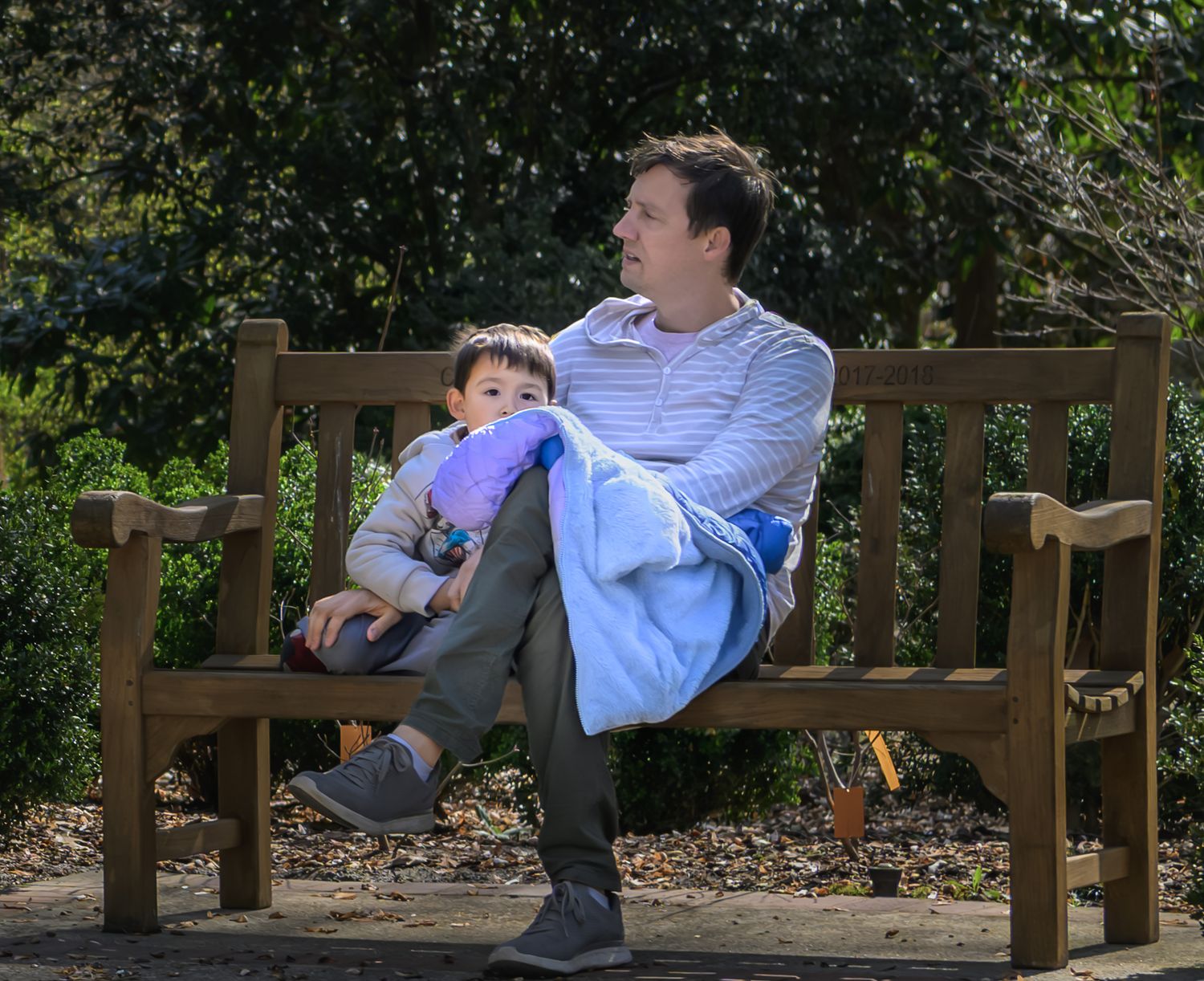 Adult and toddler sitting together on a wooden park bench outdoors on a sunny day.