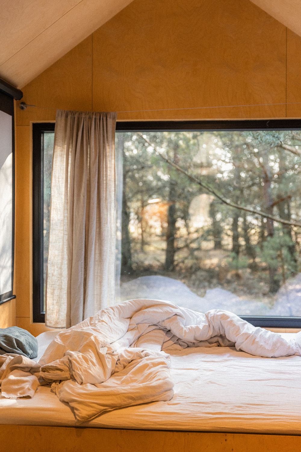 Cozy unmade bed in a tiny cabin with large windows overlooking a snowy forest, featuring warm wood walls.