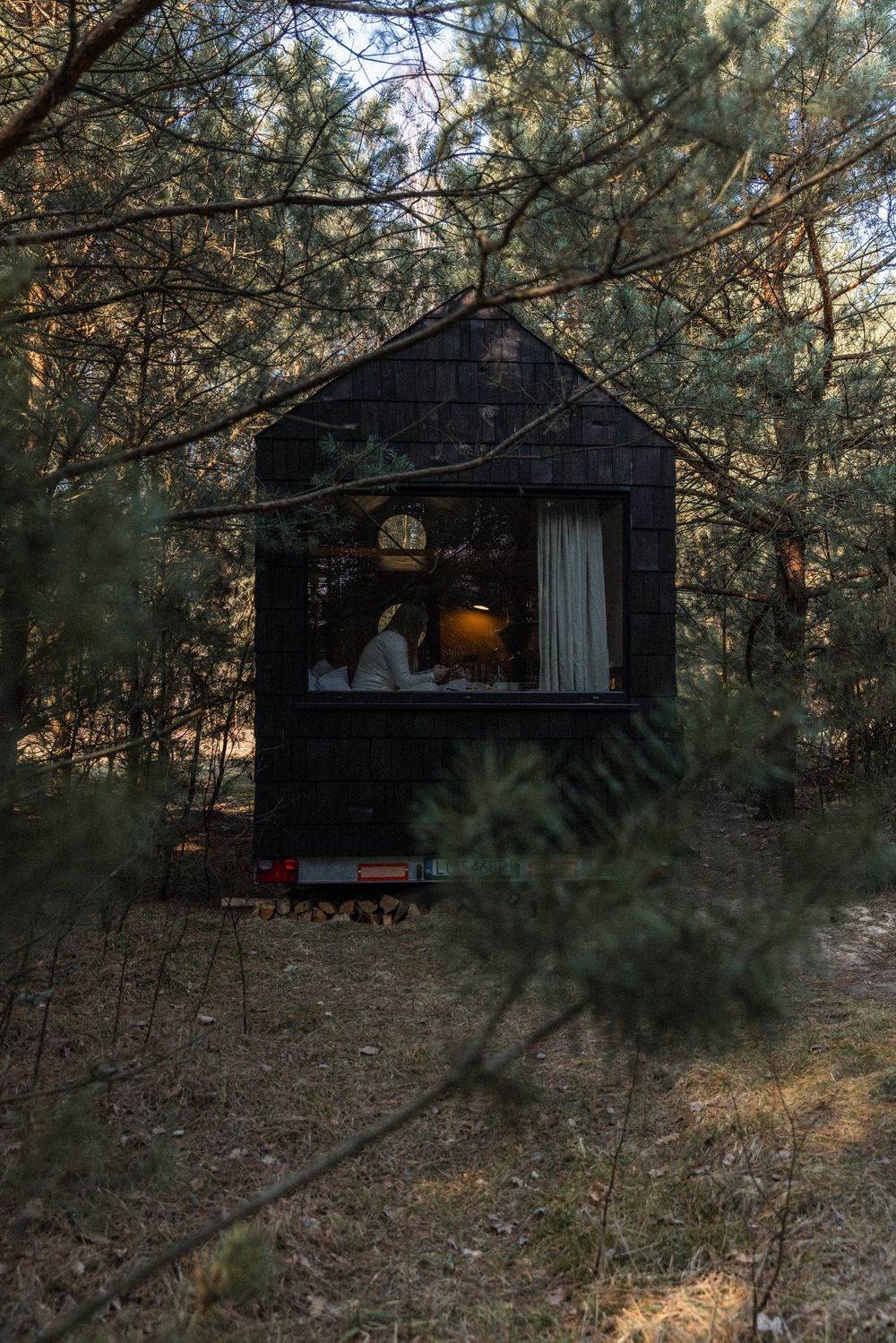 A person sits inside a cozy dark tiny cabin nestled among pine trees in a forest at dusk.