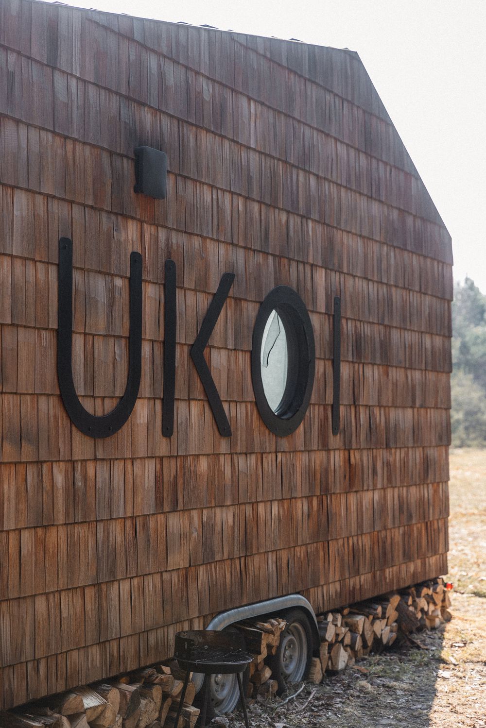 Rustic wooden tiny house on wheels with cedar shingle siding and UKOI signage, featuring a round porthole window.