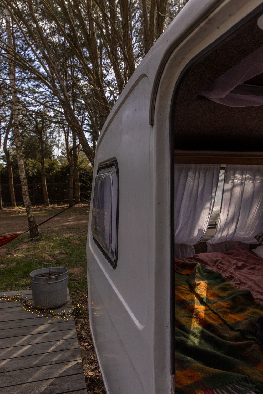 White caravan with open door revealing colorful bedding, parked on wooden deck surrounded by tall trees.