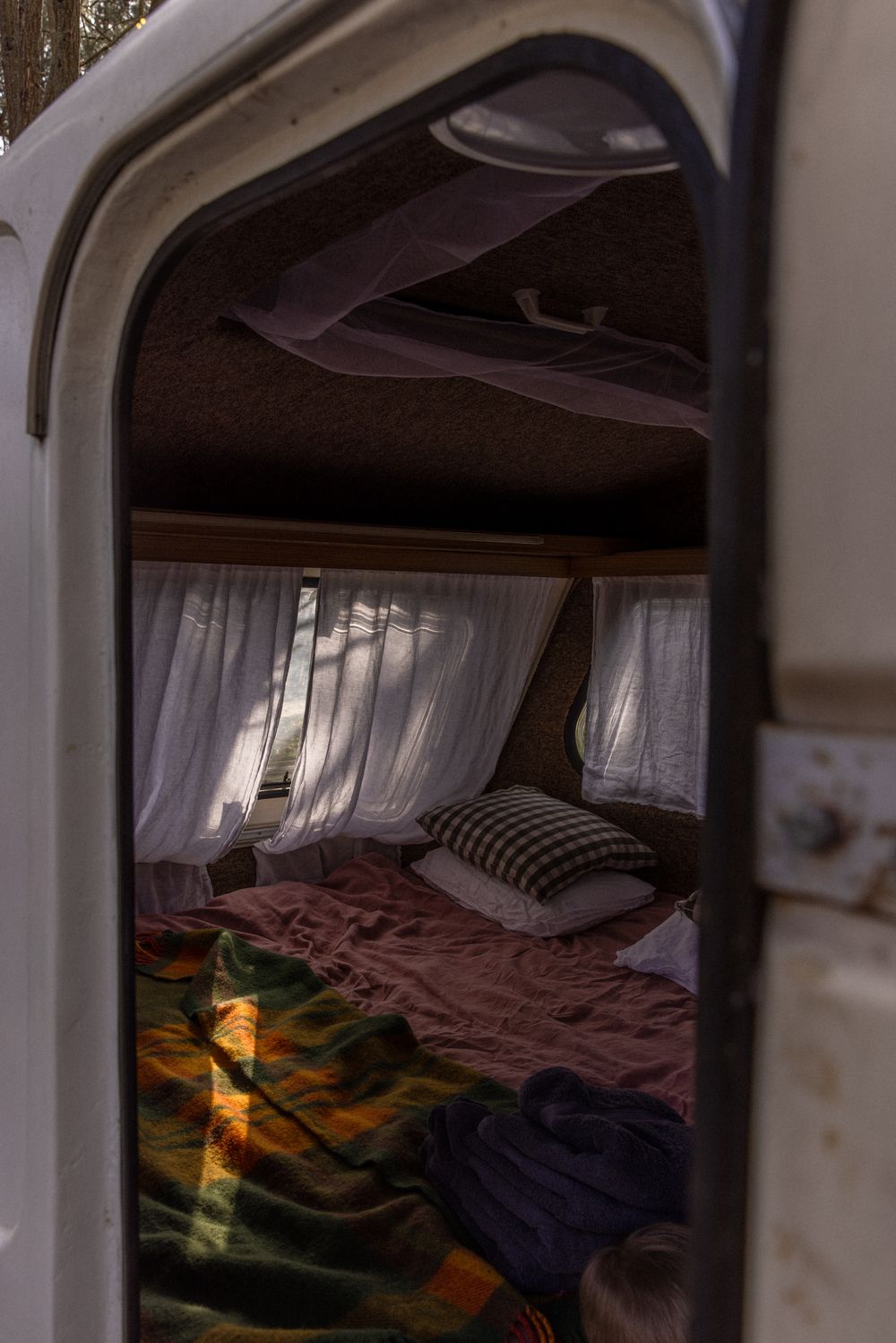 Cozy camper trailer interior with a bed covered in colorful blankets, white curtains, and warm sunlight streaming through windows.