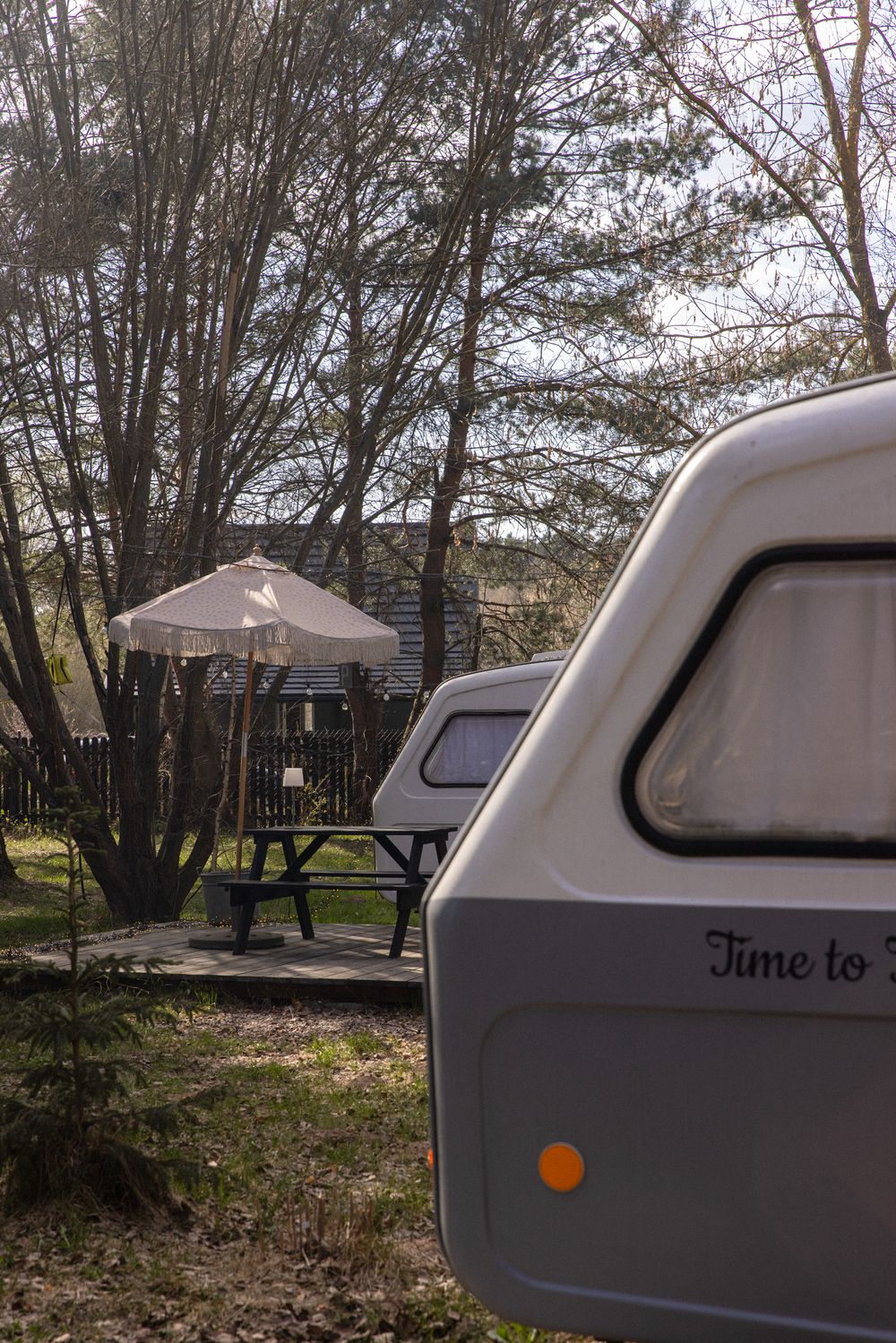 A white caravan parked among bare trees at a campsite with a picnic table and umbrella in the background.