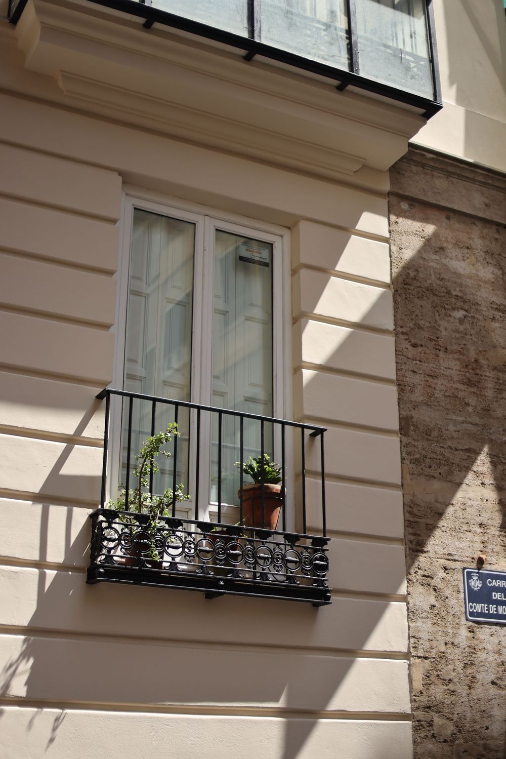 Parisian building facade with ornate black iron balcony railing, potted plants, and white French windows in warm sunlight.