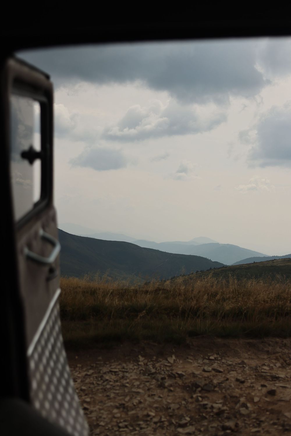 View from a vehicle door of rolling mountain hills under a cloudy sky with golden grasslands in the foreground.