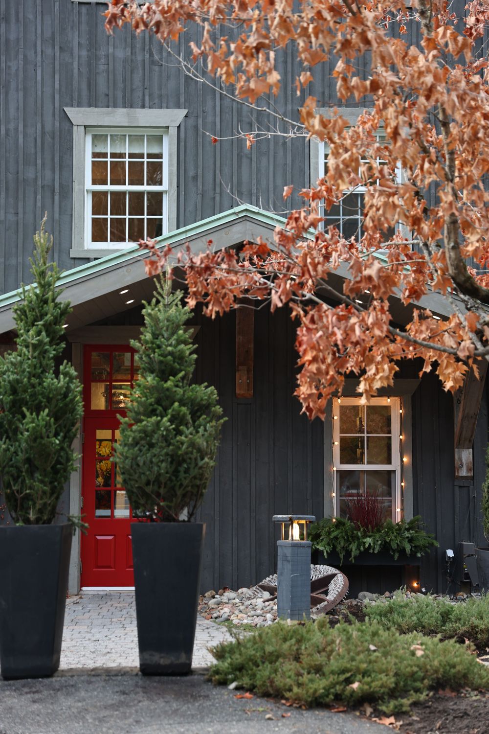 Dark gray barn-style building with red door, evergreen potted trees, autumn foliage, and a lit lantern pathway.