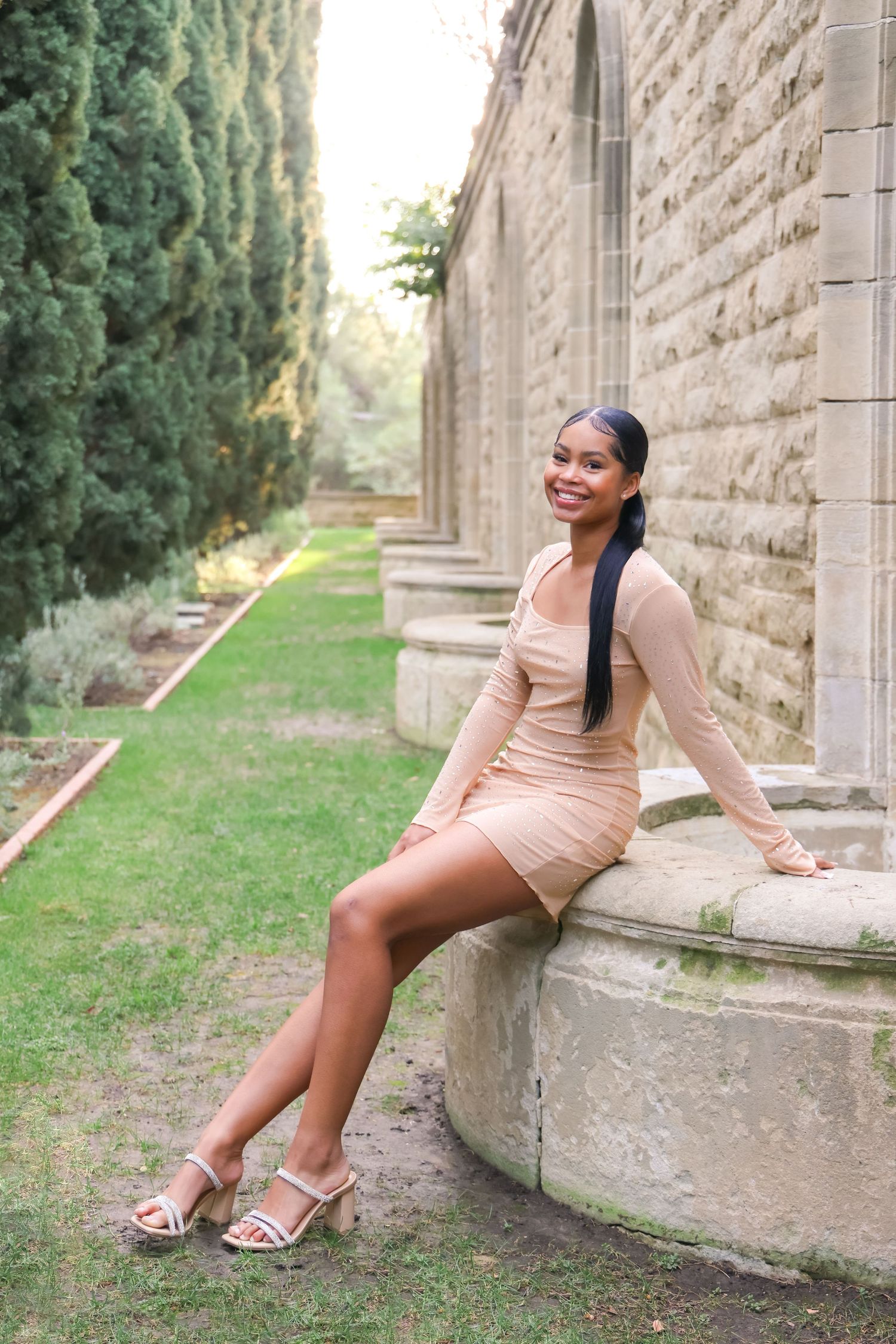 Young woman in a beige mini dress sitting on a stone ledge beside a historic building in a garden.