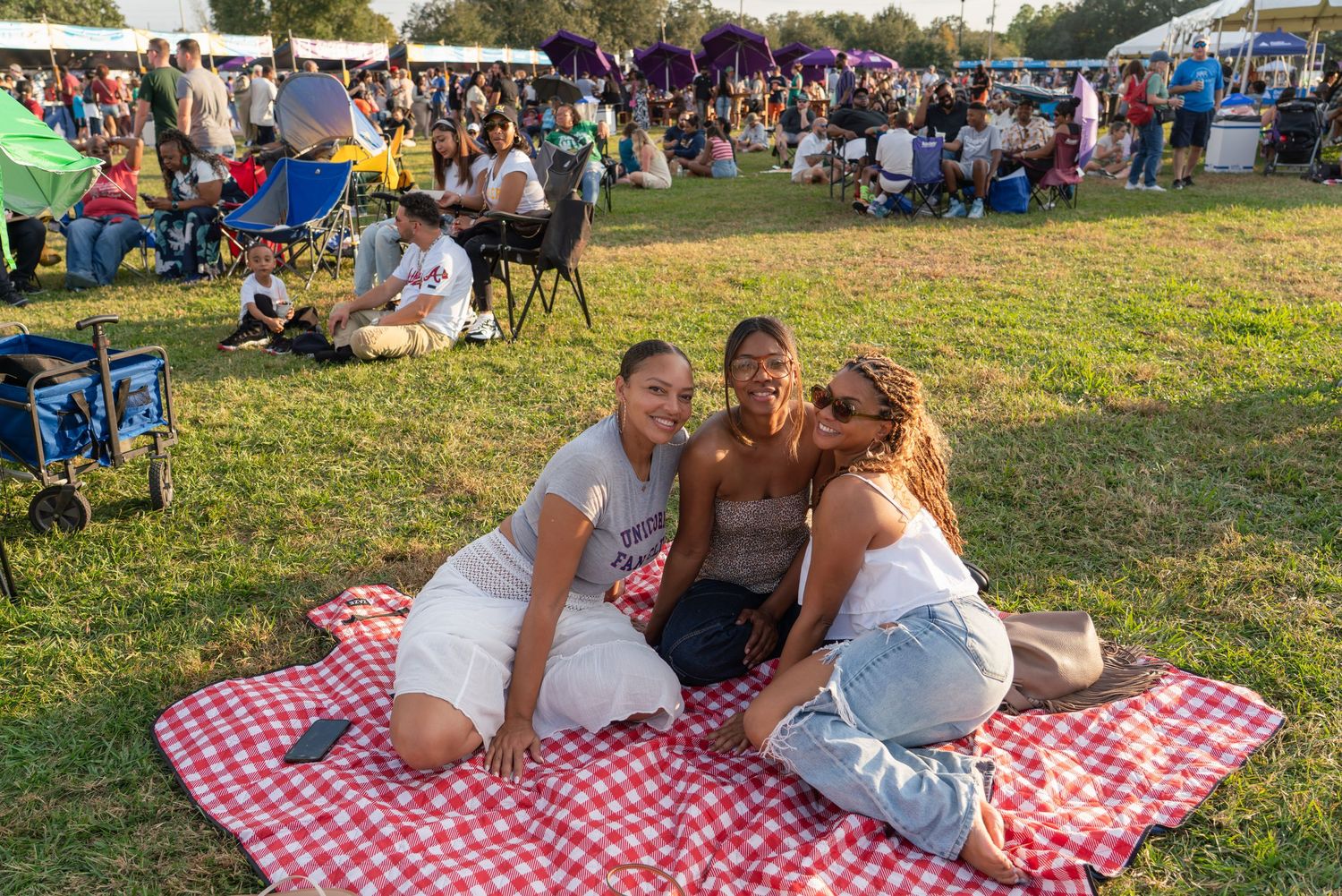 Three friends smile together on a red checkered picnic blanket at an outdoor festival on a sunny day.