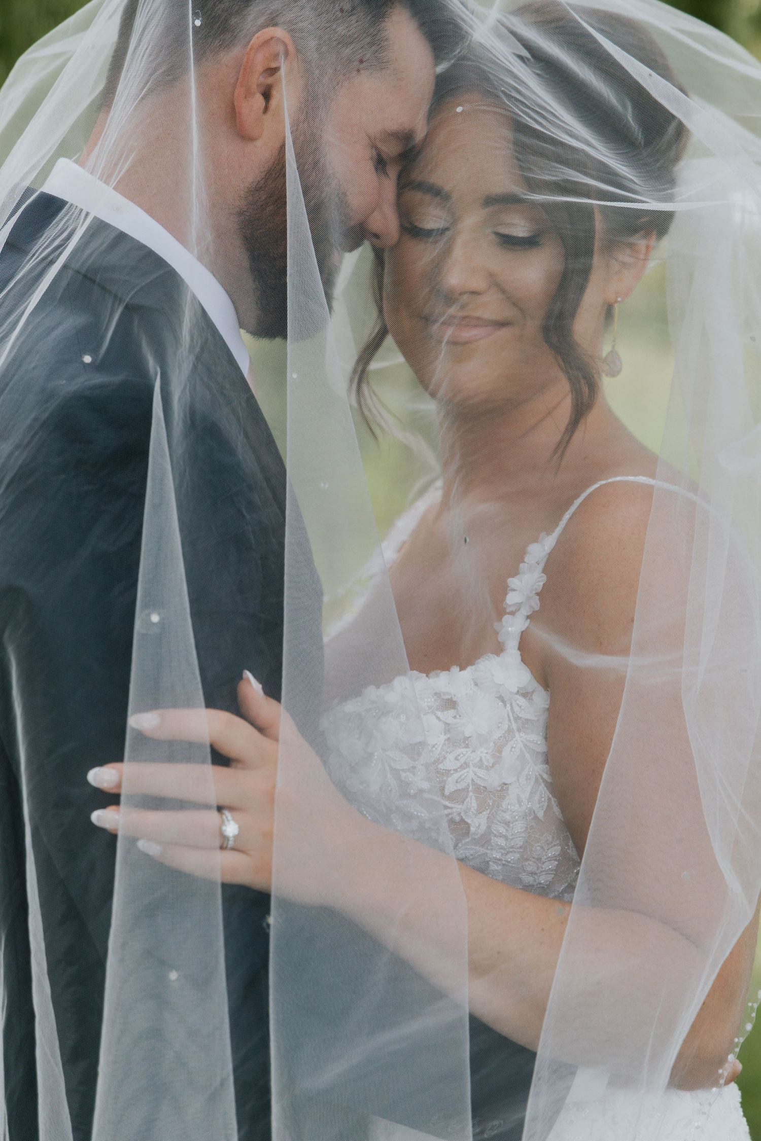 Bride and groom smiling at each other under weeping willow tree, bride holding white rose bouquet.
