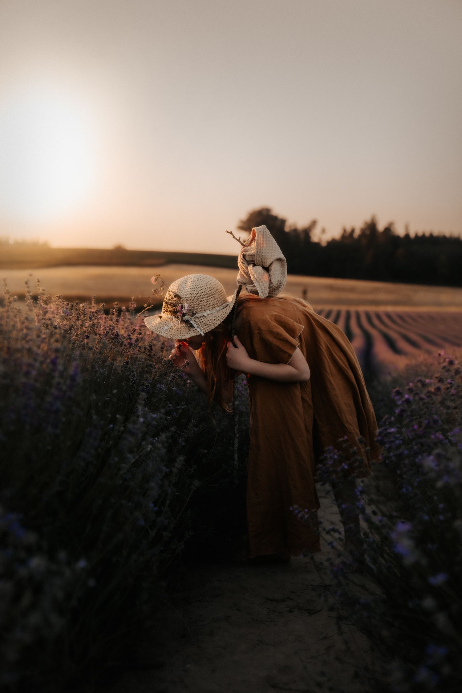 Woman in brown dress and sun hat smelling lavender flowers in a field at golden hour sunset.