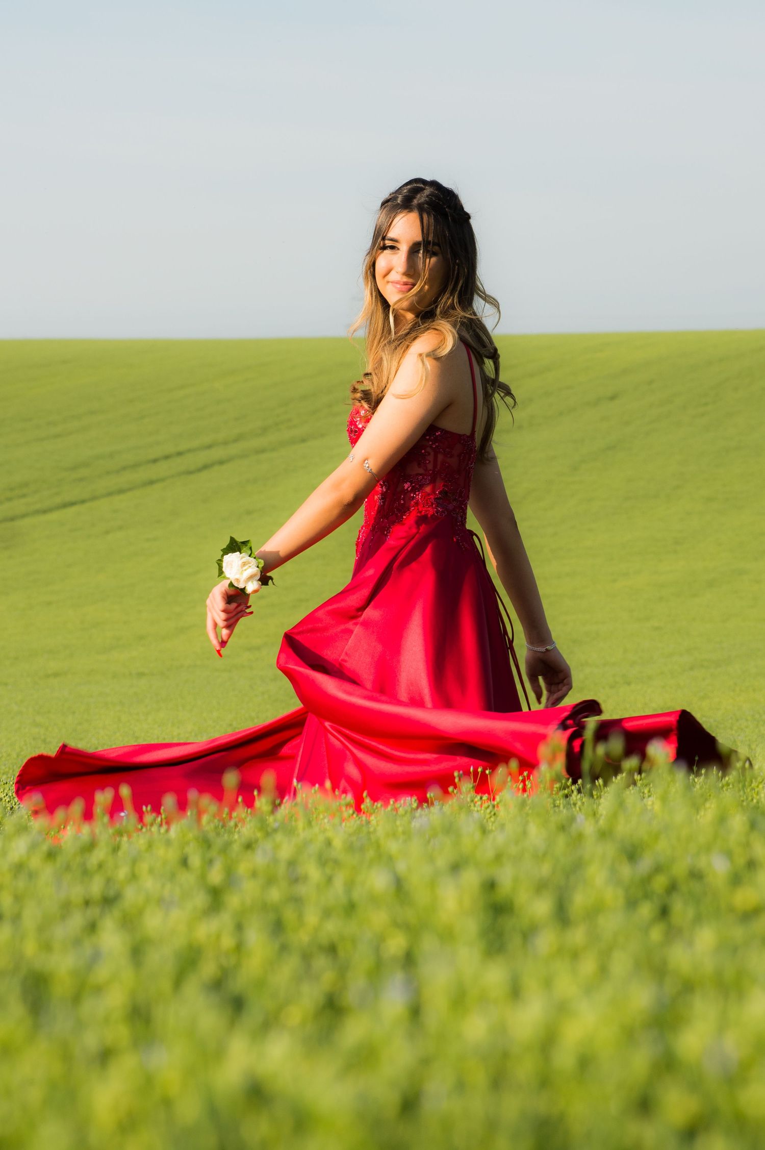 Woman in a flowing red prom gown stands in a lush green field, holding a white rose. She smiles with capturing a sense of joy and elegance.