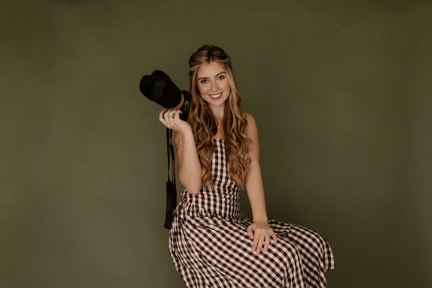 A woman in a plaid dress holds a camera, her long hair flowing, photographed against an olive green background.