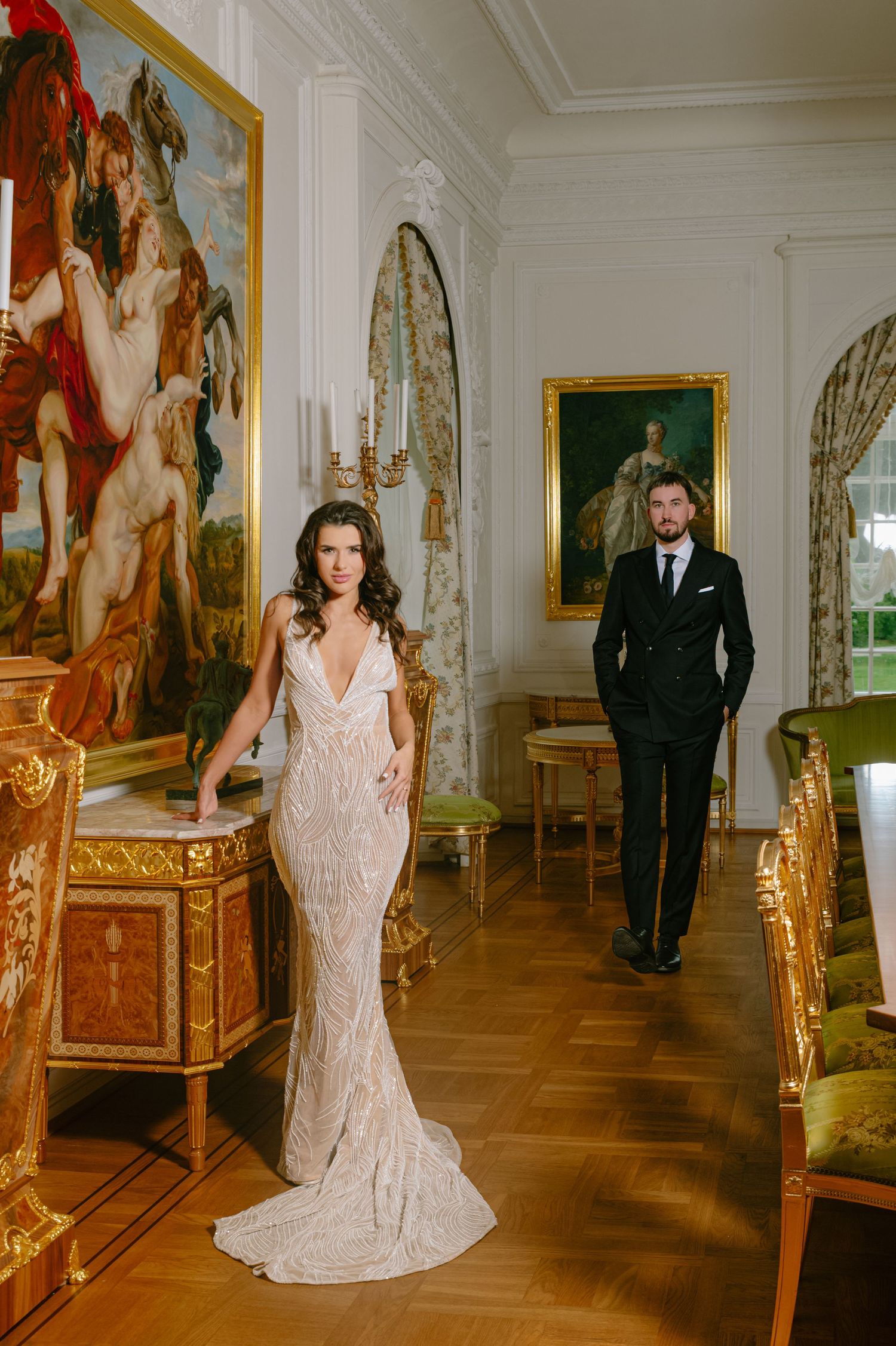 Bride in fitted lace gown and groom in black suit pose in opulent classical gallery room.
