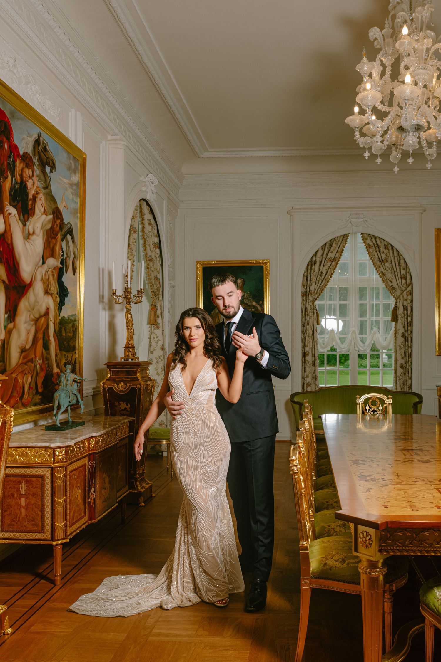 Elegant bride and groom posing in ornate ballroom with chandelier, oil paintings, and gold furnishings.
