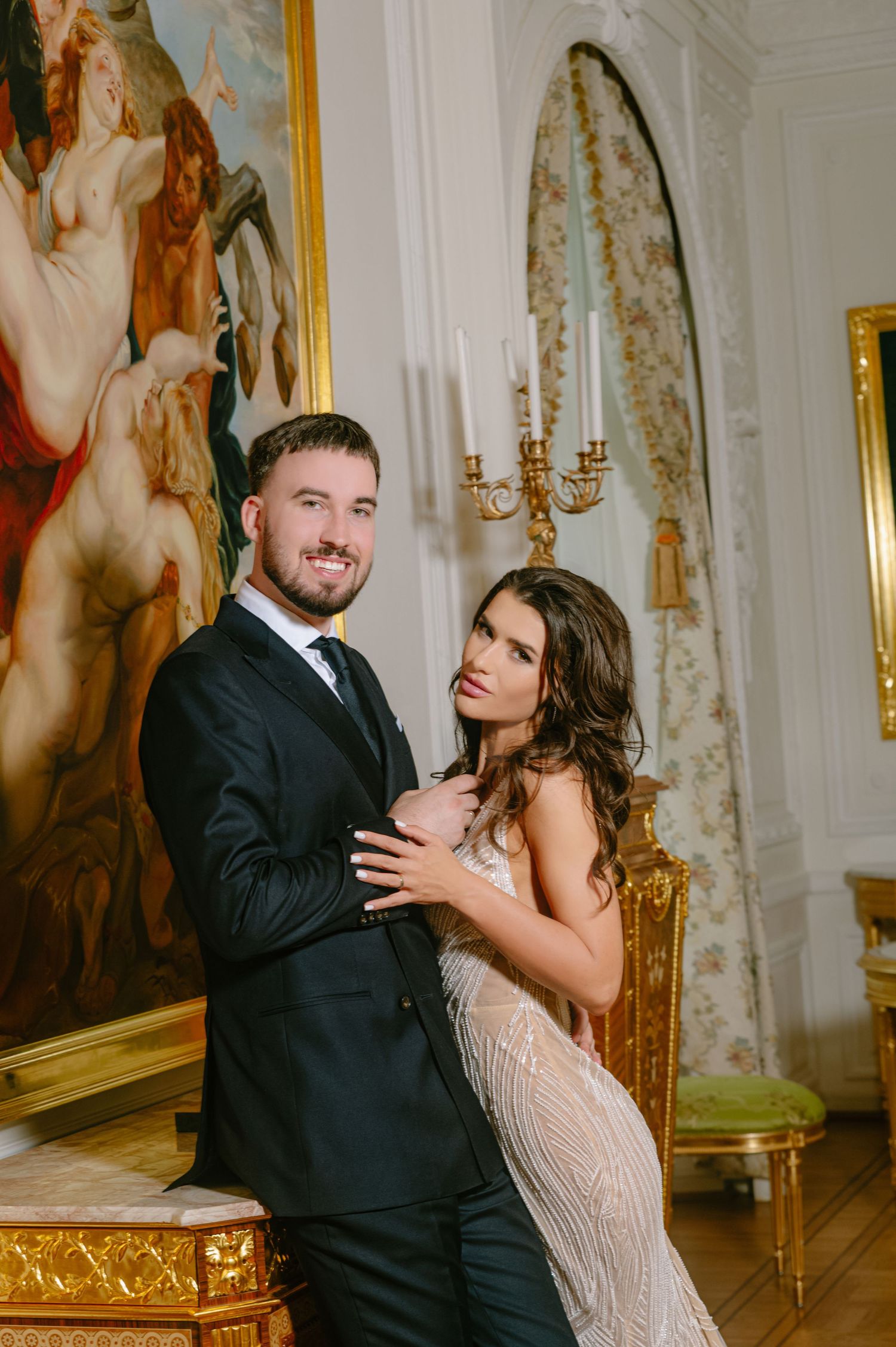 Elegant couple posing in ornate room with gold decor, man in black suit and woman in beaded gown before classic painting.