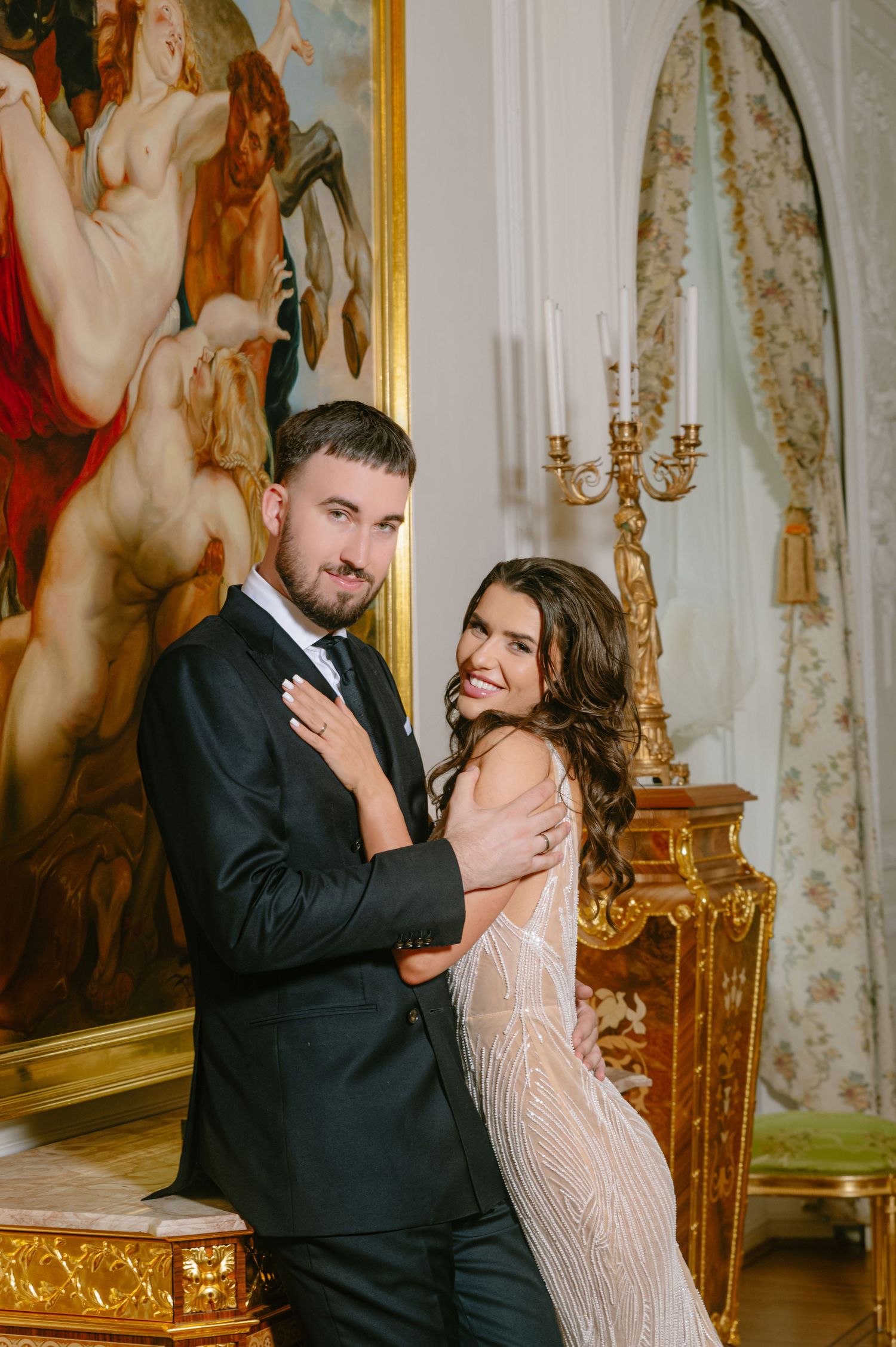 Elegant couple in wedding attire posing in ornate room with gold candelabra and classical painting backdrop.
