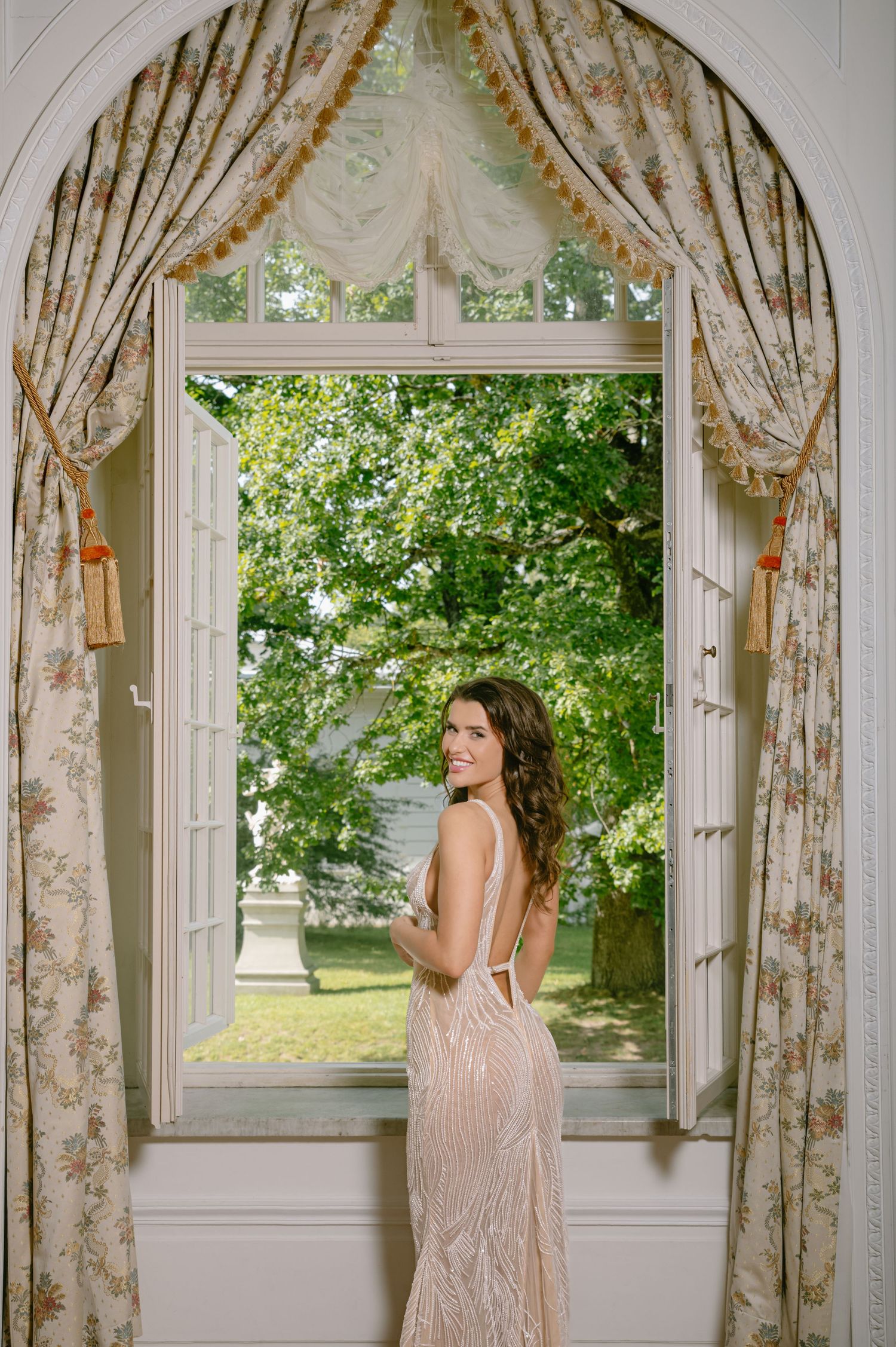 Woman in elegant backless beaded gown standing by arched window with floral curtains, lush green trees visible outside.