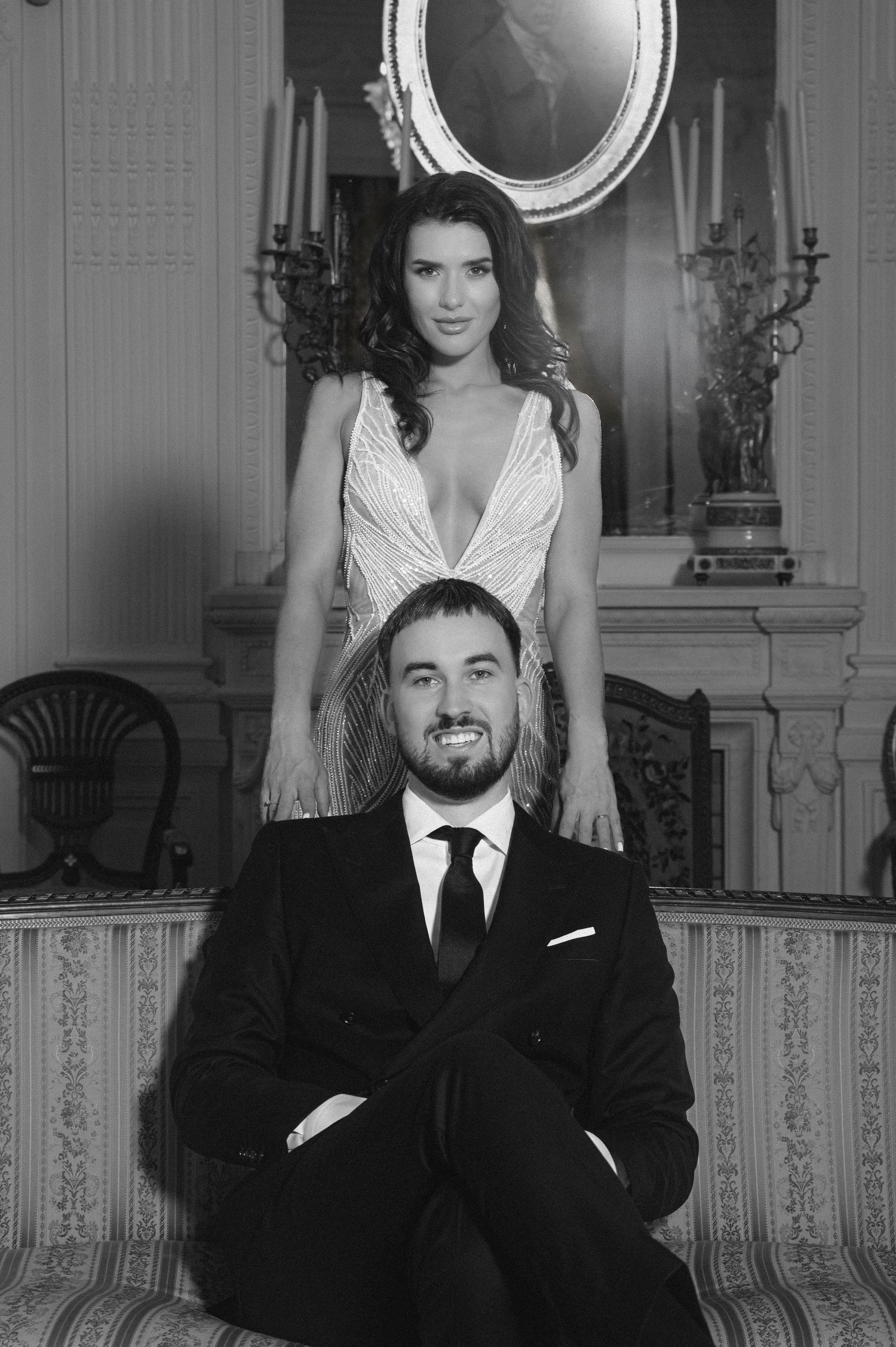 Black and white photo of a smiling bearded man in a suit seated before a woman in an elegant gown in a grand interior.