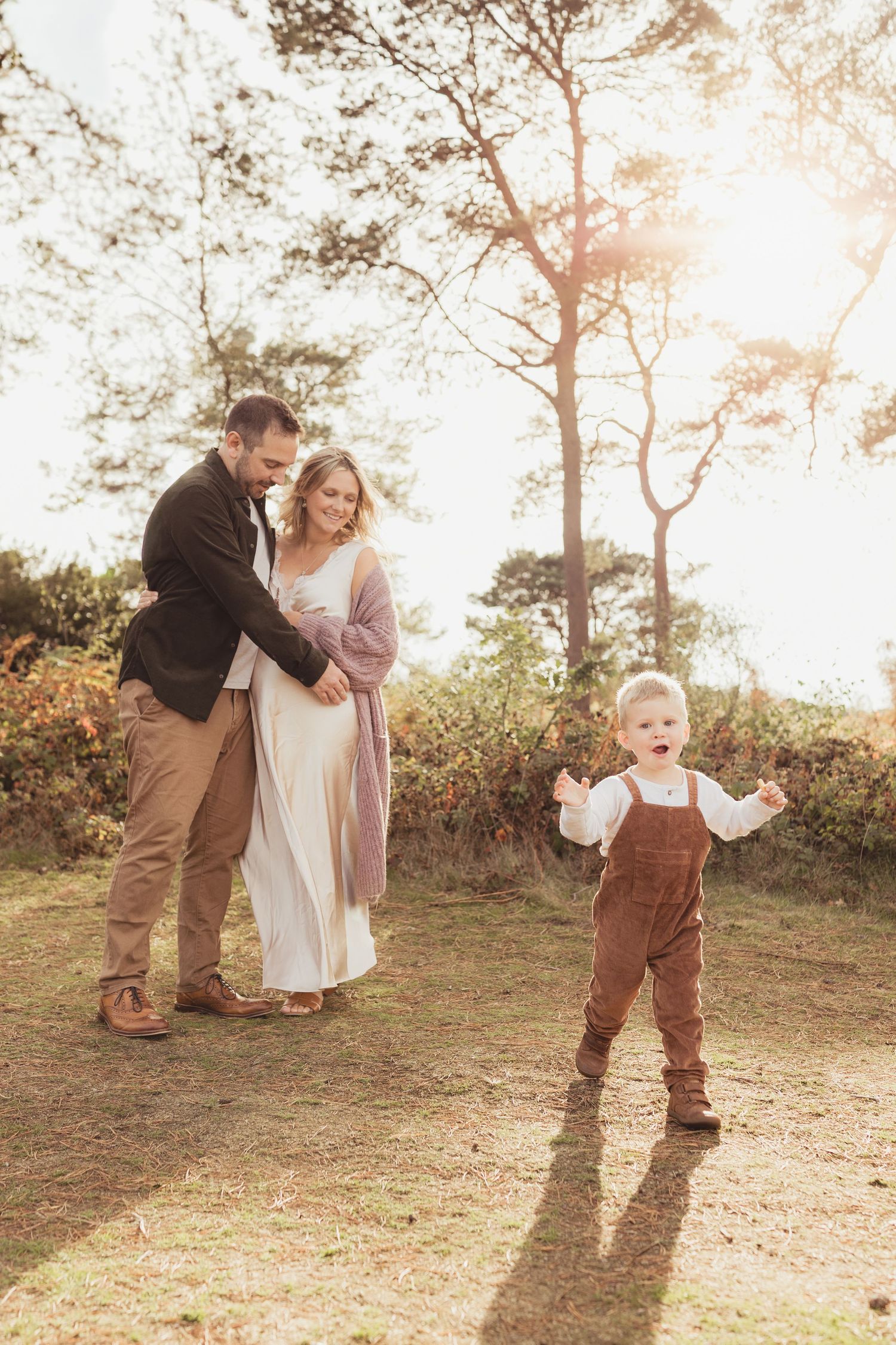 A joyful family with a toddler in brown overalls playing outdoors during a golden hour maternity photo session.
