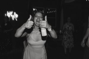 A wedding guest in a pink dress gives enthusiastic thumbs up while holding a drink at a reception.