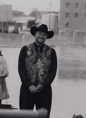 A black and white photo shows a person wearing a cowboy hat and camouflage vest in an urban setting.