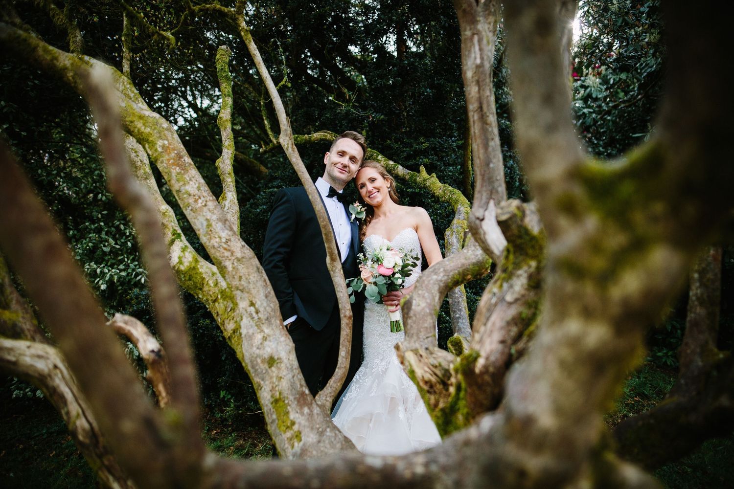 Bride and groom smiling among moss-covered trees in a lush garden, bride holding a bouquet in a fitted white gown.