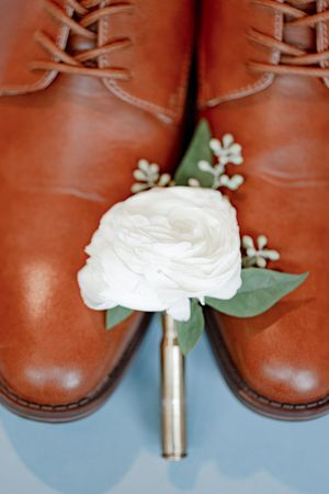 White ranunculus boutonniere with greenery resting against tan leather lace-up dress shoes on a light blue surface.