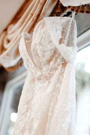 Elegant white lace wedding dress hanging near a window, showcasing intricate beading and delicate floral lace details.