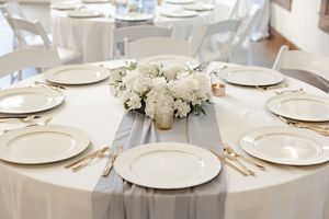 Elegant wedding reception table setting with white plates, gold cutlery, and white floral centerpiece on gray linen.