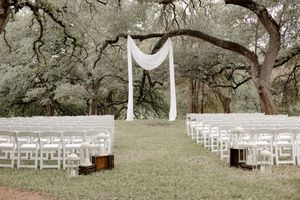 Outdoor wedding ceremony setup with white chairs, draped fabric arch, and lush oak trees in a serene garden setting.