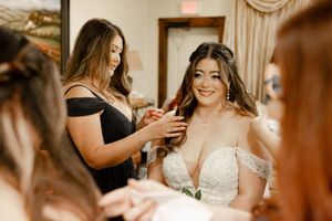 Bride in lace wedding gown gets jewelry help from bridesmaid in black dress during wedding preparation.
