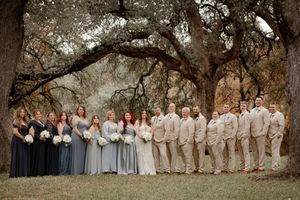 Wedding party poses under oak trees, bridesmaids in blue-gray gowns and groomsmen in tan suits.