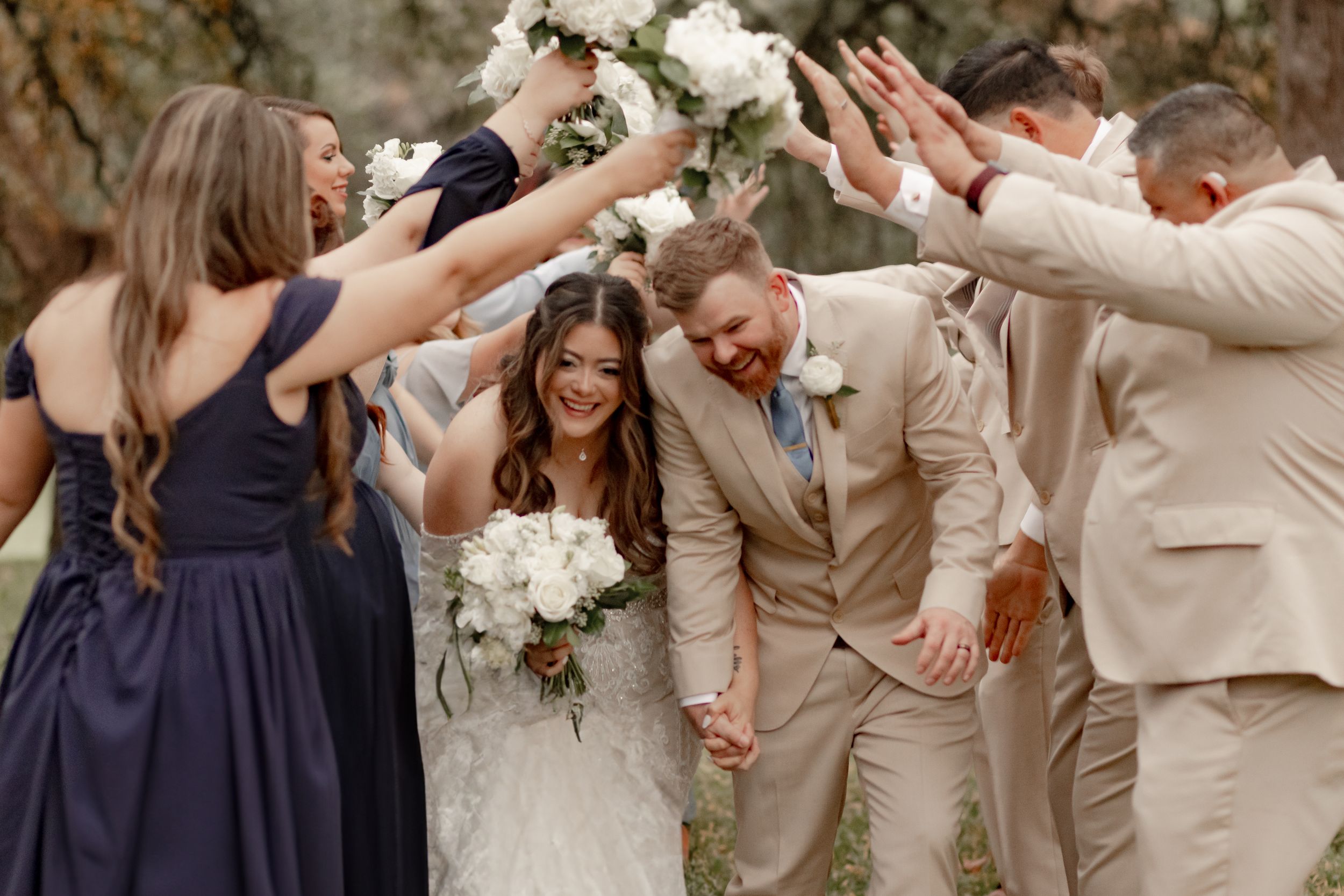 Joyful newlyweds walking hand-in-hand under raised hands of wedding party in tan and navy attire.