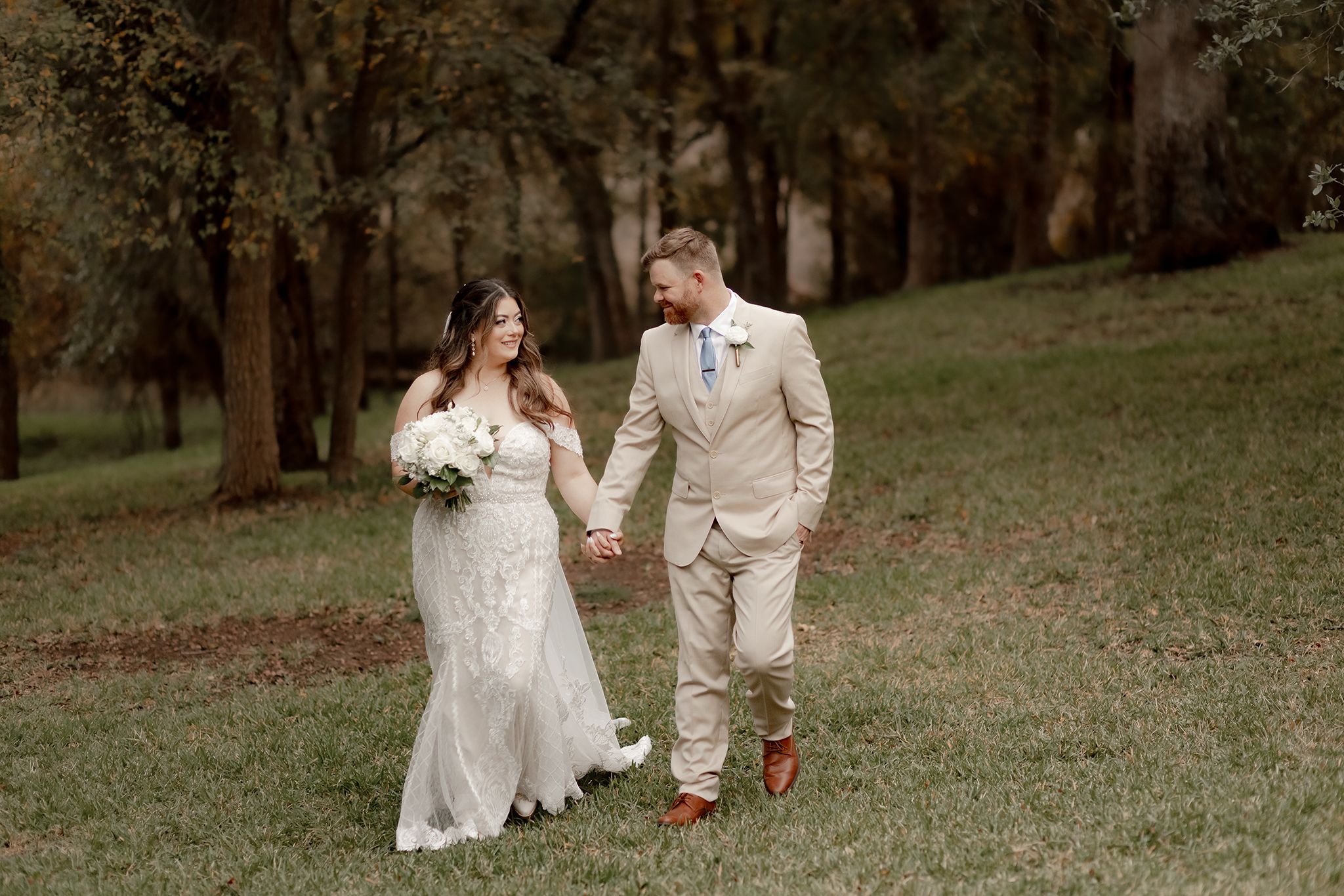 Bride and groom walk hand-in-hand through lush green grass surrounded by trees.