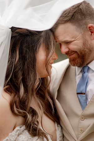 Bride and groom share an intimate forehead touch under a flowing white veil, groom wearing a tan suit with blue tie.