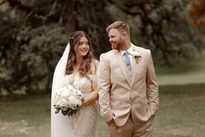Bride in white lace gown holding bouquet stands with groom in tan suit outdoors at a garden wedding.