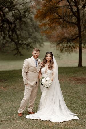 Bride in lace gown with cathedral veil holding white bouquet poses with groom in tan suit outdoors.