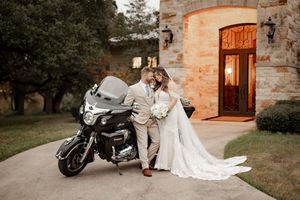 Wedding couple sharing a kiss beside a motorcycle in front of a stone venue at dusk.