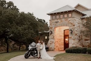 Bride and groom pose with a motorcycle in front of a stone building at their rustic Texas wedding venue.