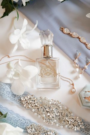 Elegant bridal flat lay with Juicy Couture perfume, rose gold jewelry, crystal sash, and white orchids on silk.