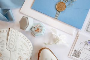 Elegant wedding flat lay with blue velvet ring box, wax seal envelope, floral invitation, and bridal shoes on white background.