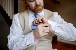 Groom with red beard adjusting cufflinks wearing beige vest, white shirt, and blue tie before wedding ceremony.