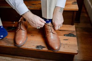 Groom tying brown leather oxford shoes with American flag socks on wooden steps before wedding ceremony.