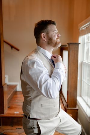 Smiling groom in tan vest adjusting his tie near a bright window while getting ready for wedding.