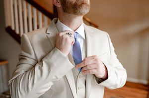 Groom in cream suit adjusting blue tie, preparing for wedding ceremony indoors.